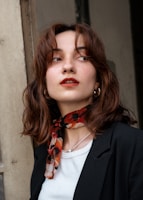 A woman with medium-length brown hair is looking off to the side. She wears a black blazer over a white shirt, and accessorizes with a red and black patterned scarf around her neck, gold hoop earrings, and a delicate necklace. Her makeup includes red lipstick.