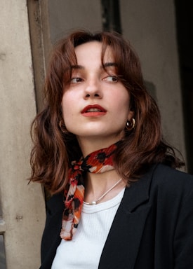 A woman with medium-length brown hair is looking off to the side. She wears a black blazer over a white shirt, and accessorizes with a red and black patterned scarf around her neck, gold hoop earrings, and a delicate necklace. Her makeup includes red lipstick.