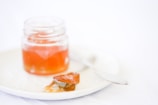 A glass jar filled with orange jelly sits on a white plate. Nearby, a spoon partially coated with the same jelly rests on the plate. The background and overall setting are minimalistic and softly lit.