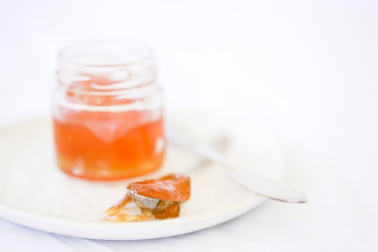 A glass jar filled with orange jelly sits on a white plate. Nearby, a spoon partially coated with the same jelly rests on the plate. The background and overall setting are minimalistic and softly lit.