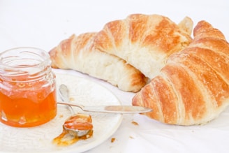 A close-up of fresh croissants placed on a white surface beside an open jar of apricot jam. The jar is on a white plate with a small spoon covered in jam lying next to it.