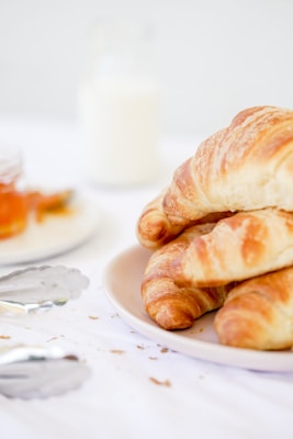 A plate with freshly baked croissants is placed on a table, along with a jar of milk in the background. The surface of the croissants has a golden brown texture, suggesting a flaky and buttery consistency. In the foreground, there is a pair of metallic tongs, hinting at a breakfast setting. The overall arrangement is minimalistic and clean, with a soft, inviting ambiance.