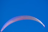 A vibrant paragliding wing is seen against a clear, cloudless sky. The wing is primarily white with hints of pink and purple hues along its edges.