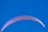 A vibrant paragliding wing is seen against a clear, cloudless sky. The wing is primarily white with hints of pink and purple hues along its edges.
