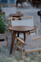 Wooden chairs with beige fabric slings surround small round wooden tables in what appears to be an outdoor cafe setting. Potted plants are placed around the area, and there's a blurred image of a person in the background, suggesting a relaxed and casual atmosphere.