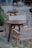 Wooden chairs with beige fabric slings surround small round wooden tables in what appears to be an outdoor cafe setting. Potted plants are placed around the area, and there's a blurred image of a person in the background, suggesting a relaxed and casual atmosphere.