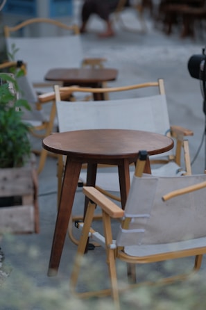 Wooden chairs with beige fabric slings surround small round wooden tables in what appears to be an outdoor cafe setting. Potted plants are placed around the area, and there's a blurred image of a person in the background, suggesting a relaxed and casual atmosphere.
