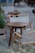 Wooden chairs with beige fabric slings surround small round wooden tables in what appears to be an outdoor cafe setting. Potted plants are placed around the area, and there's a blurred image of a person in the background, suggesting a relaxed and casual atmosphere.
