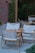 A wooden outdoor seating area consists of folding chairs with white fabric and a matching wooden table. The seating is arranged on a stone or concrete surface, bordered by white pebbles. In the background, there is a red brick wall and lush green bamboo plants providing a natural ambiance.
