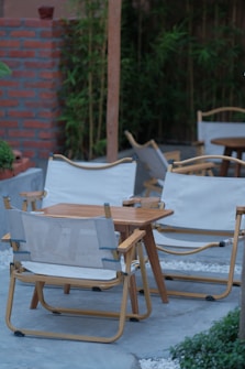 A wooden outdoor seating area consists of folding chairs with white fabric and a matching wooden table. The seating is arranged on a stone or concrete surface, bordered by white pebbles. In the background, there is a red brick wall and lush green bamboo plants providing a natural ambiance.