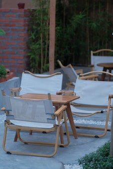 A wooden outdoor seating area consists of folding chairs with white fabric and a matching wooden table. The seating is arranged on a stone or concrete surface, bordered by white pebbles. In the background, there is a red brick wall and lush green bamboo plants providing a natural ambiance.