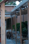 A wooden pergola with hanging light bulbs extends over a patio area. The seating consists of wooden chairs and tables, and there is greenery with large plants providing natural decoration. The setting has a relaxed and casual vibe, suggesting an outdoor dining or lounging area.