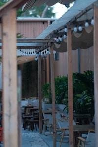 A wooden pergola with hanging light bulbs extends over a patio area. The seating consists of wooden chairs and tables, and there is greenery with large plants providing natural decoration. The setting has a relaxed and casual vibe, suggesting an outdoor dining or lounging area.