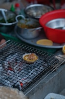 A variety of cooking ingredients and utensils are scattered around a kitchen setting. A small dish filled with food is placed on a wire mesh over hot coals, possibly grilling. There are several bowls, both metallic and plastic, some containing greens and other ingredients.