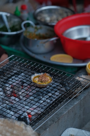 A variety of cooking ingredients and utensils are scattered around a kitchen setting. A small dish filled with food is placed on a wire mesh over hot coals, possibly grilling. There are several bowls, both metallic and plastic, some containing greens and other ingredients.