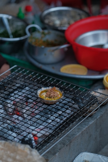 A variety of cooking ingredients and utensils are scattered around a kitchen setting. A small dish filled with food is placed on a wire mesh over hot coals, possibly grilling. There are several bowls, both metallic and plastic, some containing greens and other ingredients.