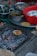A variety of cooking ingredients and utensils are scattered around a kitchen setting. A small dish filled with food is placed on a wire mesh over hot coals, possibly grilling. There are several bowls, both metallic and plastic, some containing greens and other ingredients.