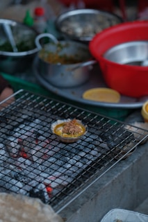 A variety of cooking ingredients and utensils are scattered around a kitchen setting. A small dish filled with food is placed on a wire mesh over hot coals, possibly grilling. There are several bowls, both metallic and plastic, some containing greens and other ingredients.