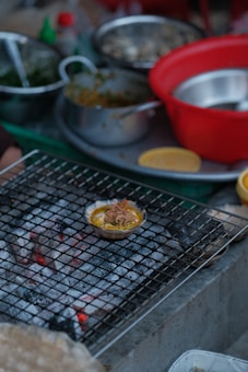 A variety of cooking ingredients and utensils are scattered around a kitchen setting. A small dish filled with food is placed on a wire mesh over hot coals, possibly grilling. There are several bowls, both metallic and plastic, some containing greens and other ingredients.