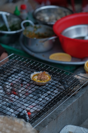 A variety of cooking ingredients and utensils are scattered around a kitchen setting. A small dish filled with food is placed on a wire mesh over hot coals, possibly grilling. There are several bowls, both metallic and plastic, some containing greens and other ingredients.