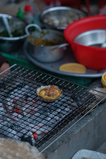 A variety of cooking ingredients and utensils are scattered around a kitchen setting. A small dish filled with food is placed on a wire mesh over hot coals, possibly grilling. There are several bowls, both metallic and plastic, some containing greens and other ingredients.