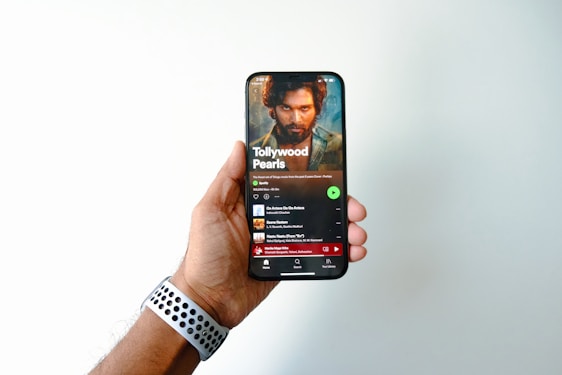 A hand is holding a smartphone displaying a music streaming app page titled 'Tollywood Pearls.' The screen shows a music playlist with a prominent image of a bearded man above the playlist title. The user's thumb is slightly visible on the side of the phone, and a watch with a perforated band is on the wrist of the hand holding the phone.