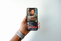 A hand is holding a smartphone displaying a music streaming app page titled 'Tollywood Pearls.' The screen shows a music playlist with a prominent image of a bearded man above the playlist title. The user's thumb is slightly visible on the side of the phone, and a watch with a perforated band is on the wrist of the hand holding the phone.