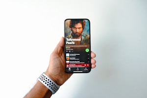A hand is holding a smartphone displaying a music streaming app page titled 'Tollywood Pearls.' The screen shows a music playlist with a prominent image of a bearded man above the playlist title. The user's thumb is slightly visible on the side of the phone, and a watch with a perforated band is on the wrist of the hand holding the phone.