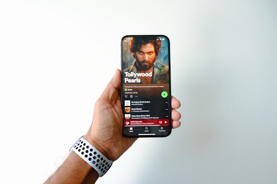 A hand is holding a smartphone displaying a music streaming app page titled 'Tollywood Pearls.' The screen shows a music playlist with a prominent image of a bearded man above the playlist title. The user's thumb is slightly visible on the side of the phone, and a watch with a perforated band is on the wrist of the hand holding the phone.