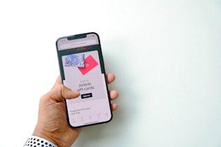 A hand is holding a smartphone displaying a screen with an advertisement for Airbnb gift cards. The screen shows a shopping option and imagery of gift cards against a bright background.