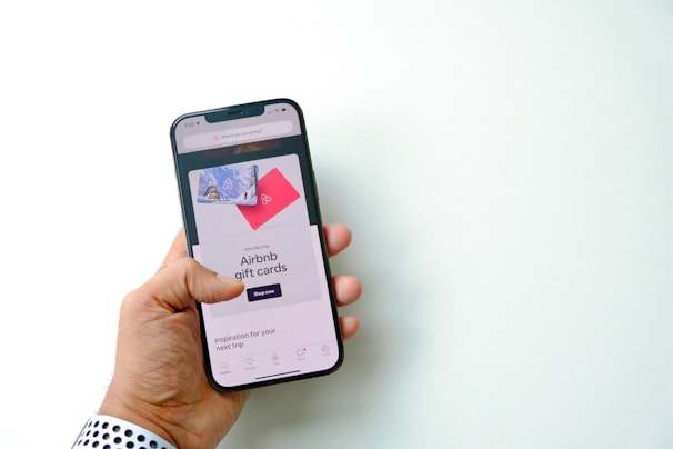 A hand is holding a smartphone displaying a screen with an advertisement for Airbnb gift cards. The screen shows a shopping option and imagery of gift cards against a bright background.
