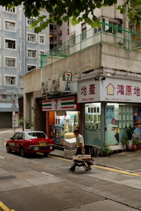 A street scene featuring a small convenience store next to a real estate office with signage in a foreign language. A man is pushing a cart filled with boxes along the street, while a red car is parked nearby. The buildings have a slightly worn urban look, with air conditioning units visible on the exterior. Trees partially frame the top of the image.