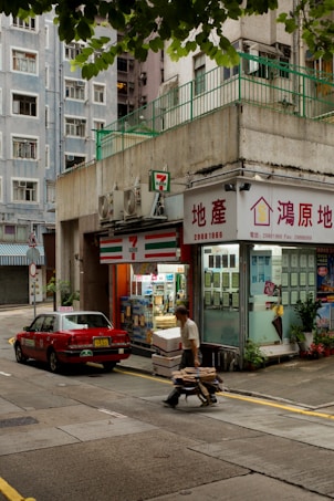 A street scene featuring a small convenience store next to a real estate office with signage in a foreign language. A man is pushing a cart filled with boxes along the street, while a red car is parked nearby. The buildings have a slightly worn urban look, with air conditioning units visible on the exterior. Trees partially frame the top of the image.