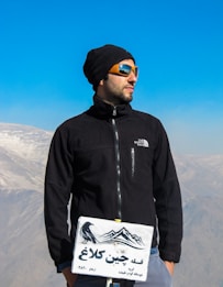 A person in outdoor gear stands confidently at a mountain summit, wearing a black jacket and orange sunglasses. In the foreground, there is a sign with a crow and mountain illustration next to text. The background showcases a clear blue sky and distant mountains partially covered in snow.