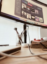 A workspace featuring a computer monitor displaying a music application with various artists' albums. Below the monitor, there is a wooden desk holding a jar with paintbrushes, a stack of books bound with twine, a white wireless earbud case, and some cables.