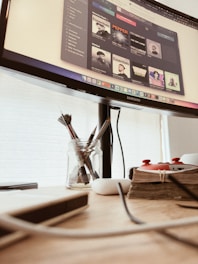 A workspace featuring a computer monitor displaying a music application with various artists' albums. Below the monitor, there is a wooden desk holding a jar with paintbrushes, a stack of books bound with twine, a white wireless earbud case, and some cables.