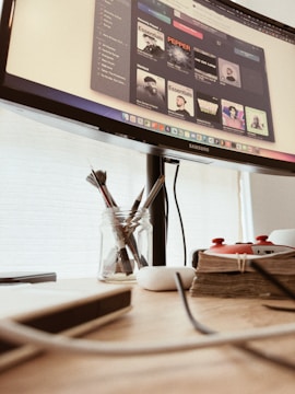A workspace featuring a computer monitor displaying a music application with various artists' albums. Below the monitor, there is a wooden desk holding a jar with paintbrushes, a stack of books bound with twine, a white wireless earbud case, and some cables.