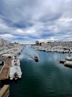 A serene harbor scene with numerous boats docked along the piers on either side of a calm waterway. Two people in a small motorized boat navigate the central waterway, creating gentle ripples as they move. The sky is overcast with thick, dramatic clouds, and buildings line the waterfront in the background.