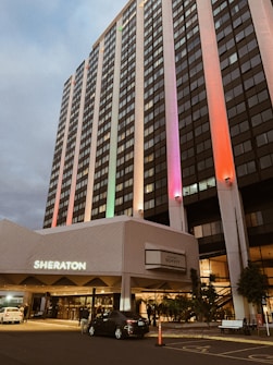 A tall, multi-story building with a prominent sign in front marked 'Sheraton.' The architecture features vertical windows and is illuminated with colorful lights on its facade. Several vehicles are visible in the foreground, with people near the entrance, suggesting the bustle of activity around a hotel.