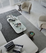 A modern living room with a minimalist design. The space features a large L-shaped sofa with striped and solid pillows, two beige armchairs, and a rectangular clear glass coffee table. The coffee table is decorated with various objects, including books, a decorative orb, and a small sculpture. The floor is covered with a large gray rug, which contrasts with the white marble flooring. Natural light fills the room from large windows.
