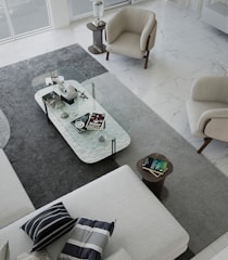 A modern living room with a minimalist design. The space features a large L-shaped sofa with striped and solid pillows, two beige armchairs, and a rectangular clear glass coffee table. The coffee table is decorated with various objects, including books, a decorative orb, and a small sculpture. The floor is covered with a large gray rug, which contrasts with the white marble flooring. Natural light fills the room from large windows.