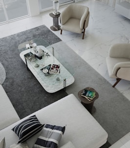A modern living room with a minimalist design. The space features a large L-shaped sofa with striped and solid pillows, two beige armchairs, and a rectangular clear glass coffee table. The coffee table is decorated with various objects, including books, a decorative orb, and a small sculpture. The floor is covered with a large gray rug, which contrasts with the white marble flooring. Natural light fills the room from large windows.