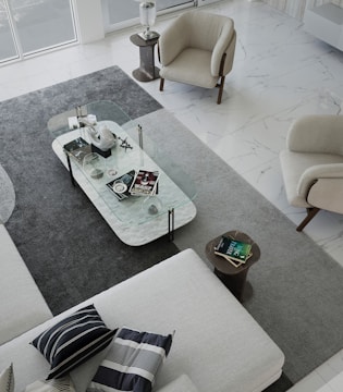 A modern living room with a minimalist design. The space features a large L-shaped sofa with striped and solid pillows, two beige armchairs, and a rectangular clear glass coffee table. The coffee table is decorated with various objects, including books, a decorative orb, and a small sculpture. The floor is covered with a large gray rug, which contrasts with the white marble flooring. Natural light fills the room from large windows.