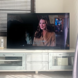 The image shows a living room with a flat-screen television displaying a scene from a movie or TV show. There is a woman with long brown hair and wearing a brown coat visible on the screen. The TV is placed on a white media console that has shelves and a cabinet. Sunlight is streaming in through partially open blinds, casting shadows on the wall and the floor.
