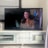 The image shows a living room with a flat-screen television displaying a scene from a movie or TV show. There is a woman with long brown hair and wearing a brown coat visible on the screen. The TV is placed on a white media console that has shelves and a cabinet. Sunlight is streaming in through partially open blinds, casting shadows on the wall and the floor.