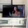 The image shows a living room with a flat-screen television displaying a scene from a movie or TV show. There is a woman with long brown hair and wearing a brown coat visible on the screen. The TV is placed on a white media console that has shelves and a cabinet. Sunlight is streaming in through partially open blinds, casting shadows on the wall and the floor.