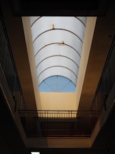A long, curved glass ceiling allows light to enter an interior space. Metal railings flank the sides, and subtle rust marks are visible on the framework supporting the glass panels. The overall structure suggests an architectural design focused on natural lighting.