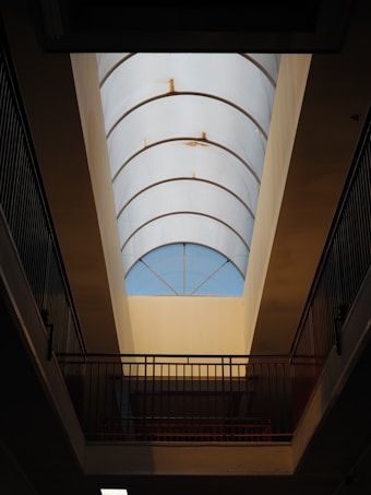 A long, curved glass ceiling allows light to enter an interior space. Metal railings flank the sides, and subtle rust marks are visible on the framework supporting the glass panels. The overall structure suggests an architectural design focused on natural lighting.