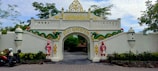 An ornate entrance gate features traditional architectural elements with decorative carvings and vibrant colors. Two statues of guards in traditional attire flank the archway. The gate is adorned with two dragon-like figures, and the name 'Puri Mataram' is inscribed in gold lettering. The setting includes trees and motorcycles parked nearby, adding a touch of everyday life to the scene.