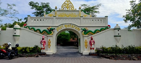 An ornate entrance gate features traditional architectural elements with decorative carvings and vibrant colors. Two statues of guards in traditional attire flank the archway. The gate is adorned with two dragon-like figures, and the name 'Puri Mataram' is inscribed in gold lettering. The setting includes trees and motorcycles parked nearby, adding a touch of everyday life to the scene.